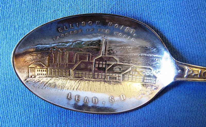 Souvenir Mining Spoon Bowl Ellison Hoist Lead SD.JPG - SOUVENIR MINING SPOON ELLISON HOIST LEAD SD - Sterling silver spoon withengraved bowl showing mine buildings and marked ELLISONHOIST LARGEST IN THE WORLD LEAD, S.D. with handle showing miner and gold pan at top, cattle below that, and a miner with pick below that, marked SOUTH DAKOTA mid-handle to bowl, 5 7/8 in. long, reverse marked Sterling  (By 1895, the Homestake Mining Company of Lead, SD recognized the need for a new, larger shaft at the mine due to its growth and expansion and the proximity of the current shafts - the Golden Star, the Golden Prospect, and the B&M - to valuable ore pockets. Construction of the Ellison Shaft on the General Ellison mineral claim began in 1895. Construction on the headframe, hoist house and crusher room got underway in 1897. The shaft was located across from the preexisting shafts on Gold Run gulch.  A 900 foot tramway was built across the gulch to haul ore from the Ellison to the Homestake Mills. The entire shaft, headframe, hoist, crushing plant and tramway were completed on January 1, 1902, costing approximately one million dollars.  The Ellison hoist was the wonder of the age.  It had hoisting engines with a capacity for 3,000 feet; crusher engines with capacity for six No. 6 Gates crushers; a compressor with capacity for 250 drills, and another compressor for the tramway and underground motors. A compressed air motor, hauling 28 steel bottom-dumping cars, containing four tons of ore each, operated between the Ellison hoist and the mills of the Homestake Mining Company, over a steel bridge 100 feet high. The Ellison shaft had three compartments. In two of them double-decked cages were operated, each deck accommodating two cars, holding a ton of ore each; the third compartment contained man-ways, air-pipes, etc.  At the start of the 20th century, the Ellison hoist and a duplicate hoist at the Anaconda copper mine in Butte, MT, along with the great hoist of the Calumet & Hecla copper mine in Michigan, which raised ore from a shaft more than one mile in depth, were the most powerful mine hoists known. The Ellison hoist had no peer on a gold mine anywhere in the world. The hoist building, housing the machinery of the Ellison equipment, was 350 feet long by 100 feet in width, and 80 feet in height over the central portion. The hoist engine was built by the Union Iron Works of San Francisco, CA.  It had two steam cylinders 30 by 72 inches and two steel reels 16 feet in diameter, which wound and payed out a flat steel cable seven inches wide by a full inch in thickness. The two cables were 3000 feet in length each.  The hoist engine was partitioned off in the big building with a glass front toward the shaft. In this compartment were placed two small air compressor engines and auxiliaries for handling and governing the movements of the great machine. The clutches, post brakes, disc brakes and reverse gear were operated by compressed air and were entirely controlled by a set of three levers and one pedal, conveniently placed on an elevated platform from which the engineer had a full view of all the machinery and the shaft. When ore was supplied sufficient to keep the hoist moving, eight tons of ore were raised to the surface every two minutes; three top men dumped the cars, feeding the ore to four No. 6 Gates crushers and returned the empties to the cage. In operation, 5,760 tons of ore were moved every 24 hours. The boiler capacity furnishing the steam power for the hoist consisted of eight Scotch marine boilers with a total capacity of 1,600 horse power. These boilers were made in the shops of W. J. Solbcrg & Son, of La Crosse, WI.  Over the next several decades, many buildings were constructed around the headframe to support operations.  On the evening of July 10, 1930, tragedy struck the Ellison Shaft.  A fire ignited from overheated pipes in the air compressor room shortly before 8 pm. Within minutes, the headframe and many smaller buildings nearby were rapidly burning.  Two men trapped in the shaft were killed. The loss of these two lives and the destruction of the Ellison Shaft were truly devastating.  The hoist, headframe and shaft were a mass of wreckage, debris, and steel beams.  The rebuild started almost immediately and was complete in February 1931.)